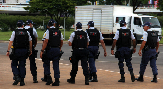 Eine Gruppe von Polizisten in Uniform, einschließlich Mützen, Hemden, Schutzwesten, Uhren, Hosen und Schuhen, geht auf einer Straße. Rechts daneben parkt ein Lastwagen in der Nähe eines Zauns, mit Gebäuden, Bäumen, Pflanzen, Pfählen und Bannern im Hintergrund und einer Brücke oben rechts.