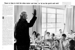 Eine Person mit Brille steht vor einer Gruppe sitzender Personen, im Hintergrund ist eine Wand mit Vorhängen und Fenstern zu sehen.