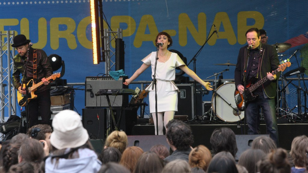 Vier Musiker auf der Bühne: Eine Frau in einem weißen Kleid singt in der Mitte, flankiert von zwei Männern, die Gitarren spielen, mit einem Publikum darunter und Lautsprechern an der Wand im Hintergrund.