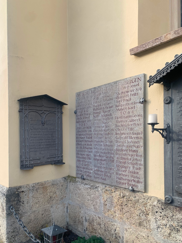 Ein Gebäude mit einer Holocaust-Gedenktafel, einem Fenster, einer Tür, einer Außenleuchte, einer Kette und umgebenden Pflanzen.
