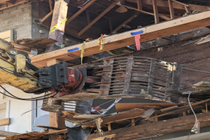 Ein Bagger arbeitet an der Seite eines baufälligen Gebäudes, das von Holzplanken und Schutt umgeben ist.