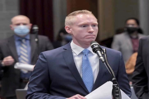 Ein Mann in Anzug und Krawatte spricht an einem Pult mit Papieren vor einem Mikrofon, während Menschen hinter ihm stehen und eine Vorhangwand zu sehen ist.