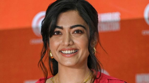 Eine Frau in einem roten Kleid mit einem eleganten Dutt und dezentem Make-up steht lächelnd da, trägt schwarze High Heels und hat ein hellgelbes Banner mit der Aufschrift 'Die besten Schönheits Looks von Sonam Kapoor beim Cannes Film Festival' im Hintergrund.
