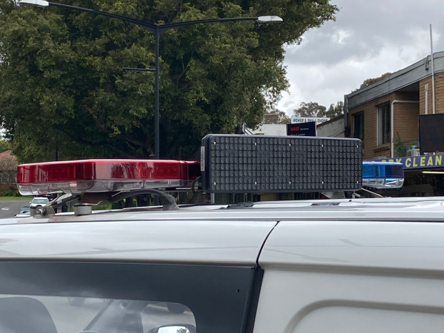 Ein Streifenwagen mit eingeschaltetem Blaulicht steht auf der Seite einer Straße, umgeben von Gebäuden, Bäumen, Pfählen und Texttafeln, mit einem bewölkten Himmel im Hintergrund, wahrscheinlich nach einem tödlichen Schulschießen in Brisbane.