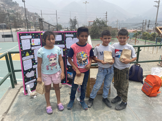 Eine Gruppe von Kindern auf einem Basketballfeld, die Papiertüten in den Händen halten, mit Tüten und Gegenständen rechts daneben und einer Tafel mit Text links daneben, vor einem Hintergrund aus Zäunen, Pfosten, Straßenlaternen, Strommasten, Bäumen, Gebäuden, Hügeln und dem Himmel.