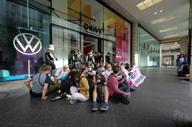 Eine Gruppe von Menschen sitzt vor einer Volkswagen-Vertretung mit Glaswänden auf dem Boden und hält ein Transparent mit Text, unter Deckenlampen.