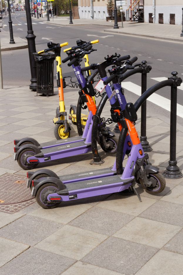 Eine Reihe von E-Scootern, die auf einer Straße geparkt sind, mit einem Mülleimer im Vordergrund, Gebäuden und Bäumen im Hintergrund und Fahrzeugen auf der Straße.