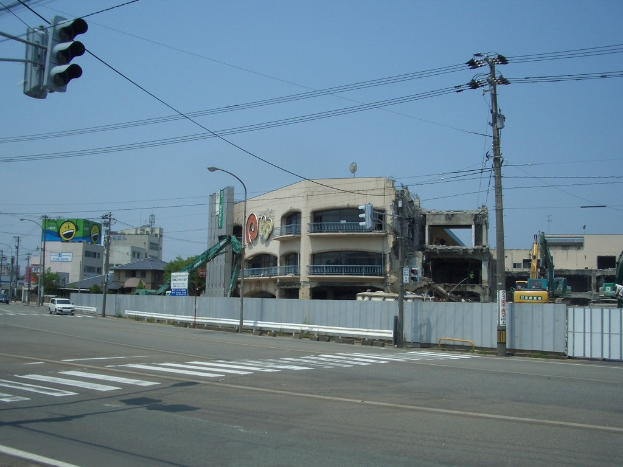 Stadtstraße mit fahrenden Fahrzeugen, Strommasten, Laternen, einer Ampel, Gebäuden, Bäumen und einem teilweise abgerissenen Gebäude im Vordergrund.