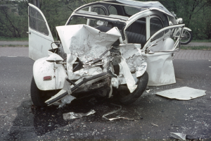 Ein Auto mit schwerer Frontsch√§digung steht am Straßenrand, mit Bäumen, einem Gebäude und einem anderen Fahrzeug im Hintergrund.