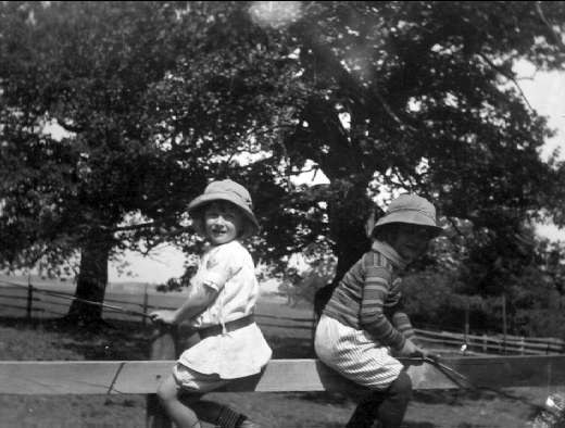Zwei junge Jungs in Hüten mit Baseballschlägern in einem grünen Park mit Bäumen und einem Zaun im Hintergrund, schwarz-weiß.
