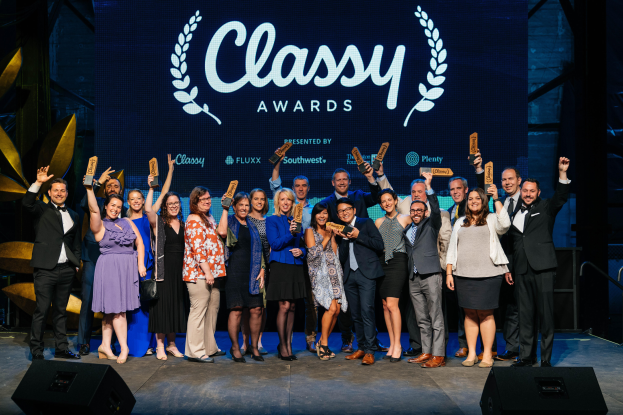 Eine Gruppe von Menschen auf einer Bühne, die lächeln und Auszeichnungen halten, mit Lautsprechern unten und einem Bildschirm im Hintergrund mit der Aufschrift "Classy Awards 2019".