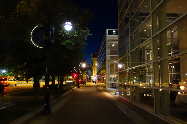 Stadtstraße bei Nacht mit beleuchteter Straße, fahrenden Fahrzeugen, Bäumen an den Seiten und Gebäuden im Hintergrund unter einem dunklen Himmel.