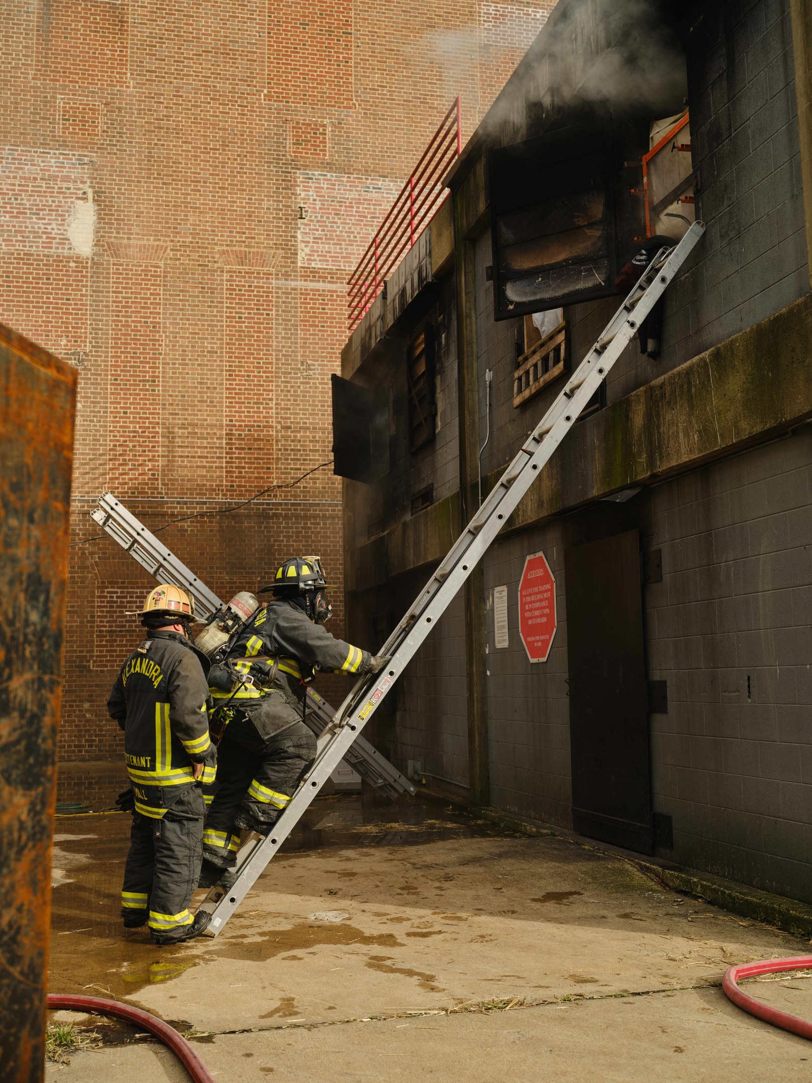 Feuerwehrleute in Helmen klettern eine Leiter hinauf zu einem raucherfüllten Gebäude, mit einem Metallobjekt links und Rohren am Boden darunter.