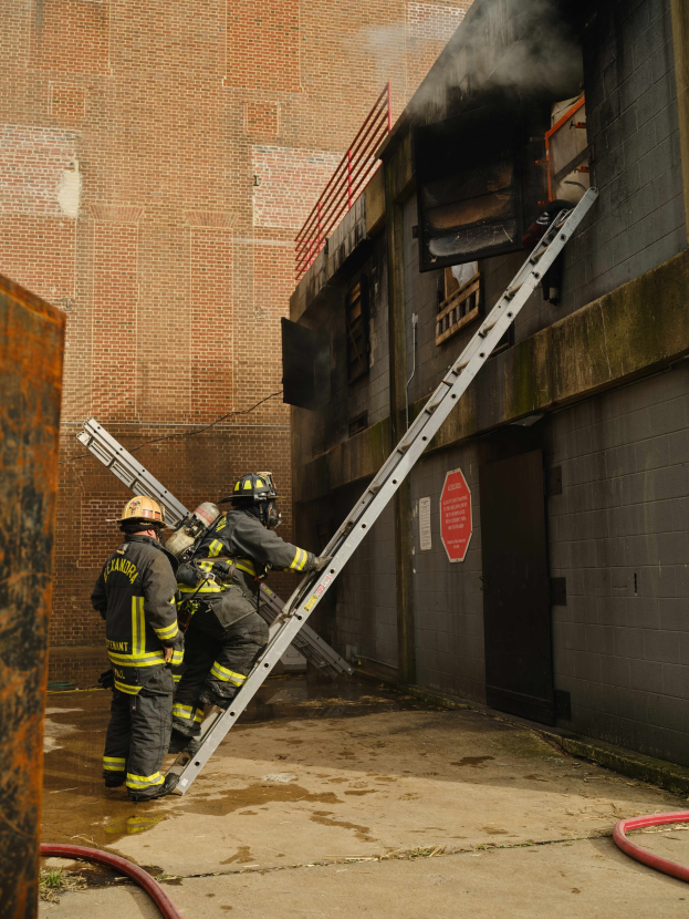 Feuerwehrleute in Helmen klettern eine Leiter hinauf zu einem raucherfüllten Gebäude, mit einem Metallobjekt links und Rohren am Boden darunter.