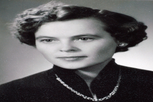 Ein Schwarz-Weiß-Foto einer Frau mit ernstem Gesichtsausdruck, deren Haar zurückgebunden ist und die ein hochgeschlossenes, langärmliges Kleid und eine Kette trägt.