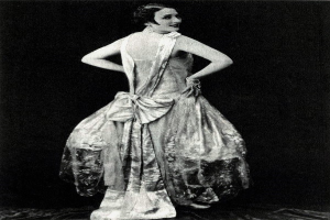 Eine Frau in einem Kleid im Stil der 1920er Jahre mit hohem Kragen, langen Ärmeln und einem weiten Rock steht selbstbewusst, ihr Haar ist elegant frisiert und das Make-up natürlich.