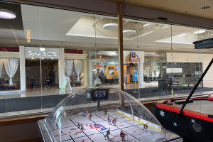 Eine Eishalle in einem Einkaufszentrum, umgeben von Glaswänden mit sichtbaren Menschen, Möbeln und Beschriftungen von außen und einem Air-Hockey-Tisch auf der rechten Seite.