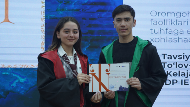 Ein Mann und eine Frau in Akademikerkleidung stehen nebeneinander und halten jeweils ein Zertifikat in der Hand, mit einem Bildschirm im Hintergrund.