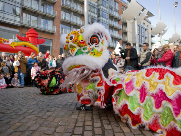 Ein lebendiges chinesisches Neujahrsfest in Amsterdam mit einem Löwen tanzen im Vordergrund mit einer Menge von Menschen, einige halten Kameras, und Gebäude, Laternenmasten und einen klaren blauen Himmel im Hintergrund.