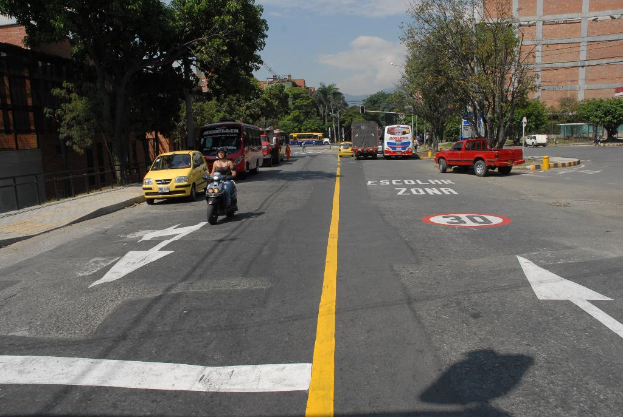 Eine Straßenszene mit Gebäuden, Bäumen, fahrenden Fahrzeugen, Schildern, Verkehrsampeln an Pfählen, einem blauen bewölkten Himmel und Warnschildern auf der Straße.
