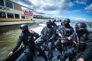 Eine Gruppe von Polizisten in schwarzen Uniformen und Helmen, einige bewaffnet, fährt auf einem Boot im Wasser mit einer Brücke, Bäumen, Gebäuden und einem klaren blauen Himmel im Hintergrund.