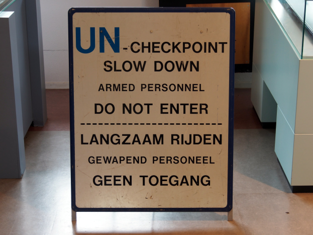 Schild auf dem Boden mit der Aufschrift "UN-Checkpoint Verlangsame bewaffnete Personen - Betreten verboten" mit einer Glasplatte und Gegenständen rechts daneben und einer Wand mit einem Gegenstand im Hintergrund.