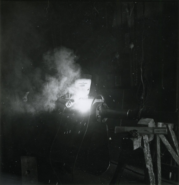 Schwarz-weißes Foto eines Schweißers bei der Arbeit an einem Metallstück, mit Rauch aus der Schweißmaschine und diversen Gegenständen im Hintergrund.