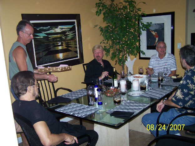 Eine Gruppe von Menschen sitzt an einem Esstisch mit Weingläsern, Flaschen und einer Schüssel mit Essen, ein Mann serviert, Fotorahmen an der Wand und eine Pflanze auf der Seite.