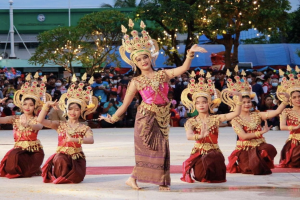 Eine Gruppe von Frauen in traditioneller thailändischer Kleidung tanzt vor einem sitzenden und stehenden Publikum im Freien, mit Bäumen, Lichtern, Pfählen, einem Gebäude und einem klaren blauen Himmel im Hintergrund.