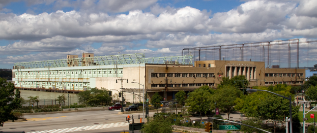 Eine Straße mit Fahrzeugen, Bäumen und Straßenlaternen entlang einer Wasserfläche mit einem großen Gebäude im Hintergrund.