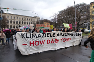 Eine Gruppe von Menschen marschiert mit einem Banner, auf dem "Klimaziele Abschaffen - Wie wagst du es?" steht, die Straße entlang. Einige halten Schirme und einer verwendet ein Megafon, vor dem Hintergrund von Laternenmasten, Schildern, Bäumen, Gebäuden mit Fenstern, einem Kran und einem klaren blauen Himmel.