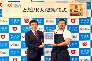 Zwei lachende Männer mit einem Zertifikat, mit einem Banner im Hintergrund, das "Toda City Wins PR Award" lautet und ein Foto einer Person zeigt.