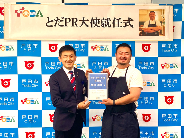 Zwei lachende Männer mit einem Zertifikat, mit einem Banner im Hintergrund, das "Toda City Wins PR Award" lautet und ein Foto einer Person zeigt.