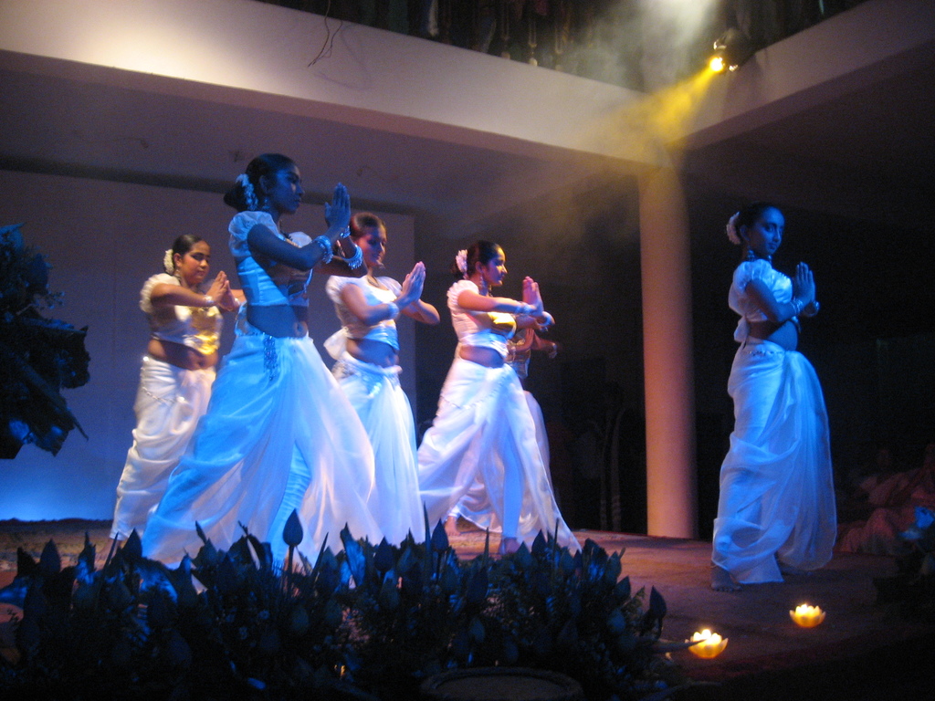 Eine Gruppe von Menschen tanzt auf einer Bühne mit Pflanzen davor und eine weitere Gruppe schaut zu.