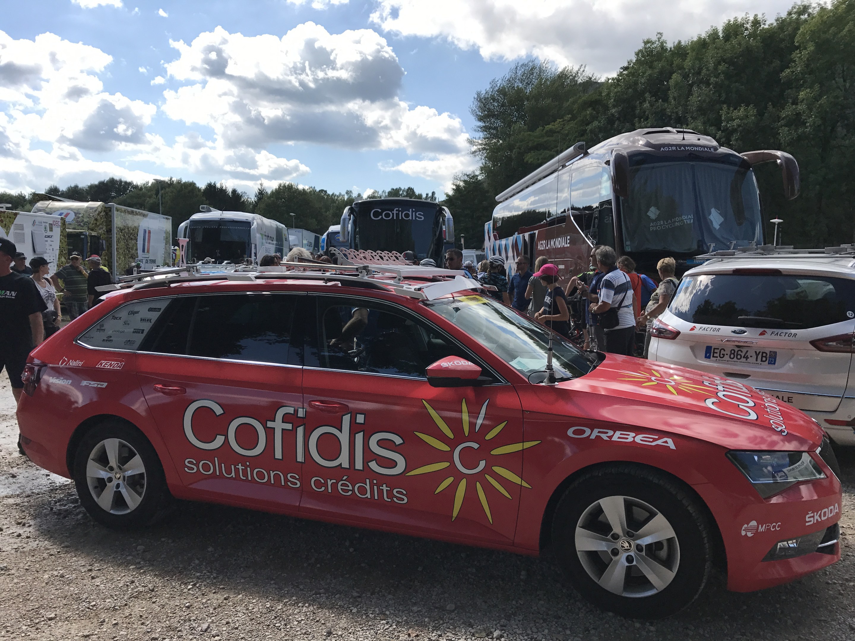 Gruppe von Menschen, die um ein rotes Cofidis Solutions Credit Team Auto in einem baumbestandenen Parkplatz unter einem klaren blauen Himmel stehen.