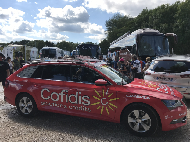 Gruppe von Menschen, die um ein rotes Cofidis Solutions Credit Team Auto in einem baumbestandenen Parkplatz unter einem klaren blauen Himmel stehen.