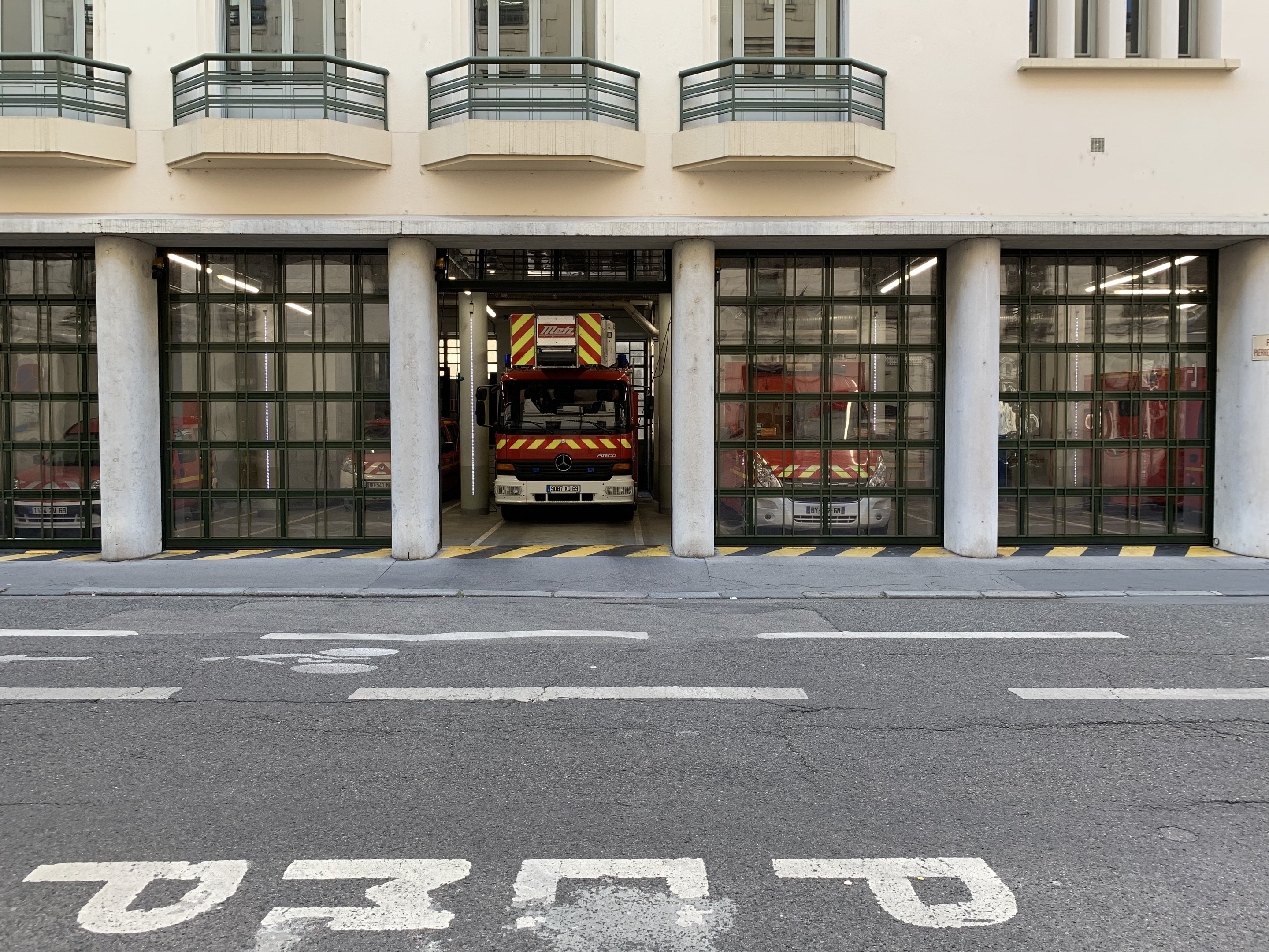 Feuerwache mit Feuerwehrauto davor, mit Fenstern, Geländern, Säulen und Glastüren, mit Straße unten.