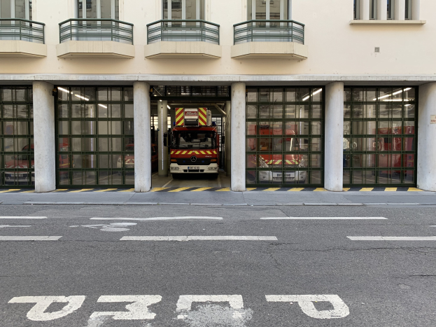 Feuerwache mit Feuerwehrauto davor, mit Fenstern, Geländern, Säulen und Glastüren, mit Straße unten.