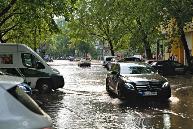 Überschwemmte Straße in Berlin, Deutschland mit Autos, die durch Wasser fahren, Fußgängern auf dem Gehweg, Bäumen am Straßenrand, Gebäuden im Hintergrund und Schildern und Pfosten, die teilweise untergetaucht sind.