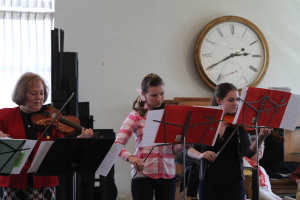 Drei Frauen auf der Bühne mit Geigen spielend, mit Notenpulten und Noten vor sich, während ein Zuschauer zuhört; eine Uhr ist an der weißen Wand im Hintergrund angebracht, und ein weißer Vorhang ist links sichtbar.