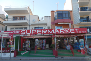 Ein Supermarkt an einer Straßenecke mit verschiedenen Geschäften, Schildern und Gebäuden im Hintergrund unter einem sichtbaren Himmel.