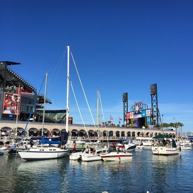 Viele Boote auf dem Wasser im Vordergrund, mit einer Brücke mit mehreren Bögen, Fahnen, Bannern, Gebäuden, Pfählen, Bäumen und einem bewölkten Himmel im Hintergrund.