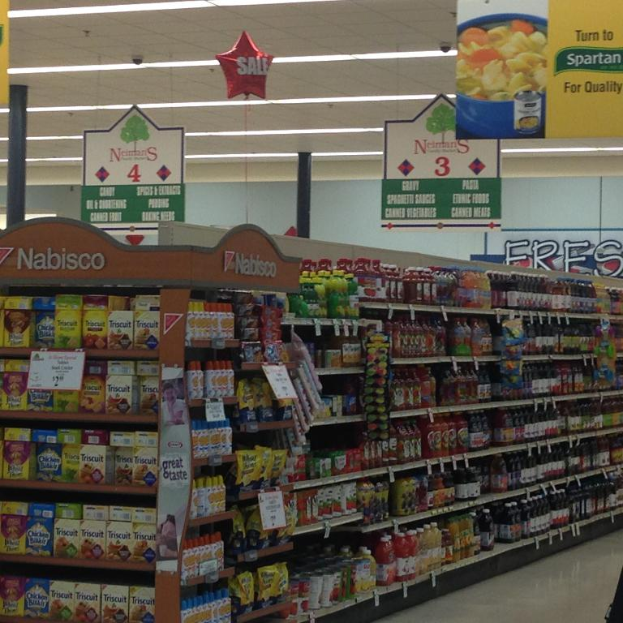Ein Supermarkt-Innenraum mit Regalen voller Lebensmittel, Deckenleuchten und von der Decke hängenden Schildern.
