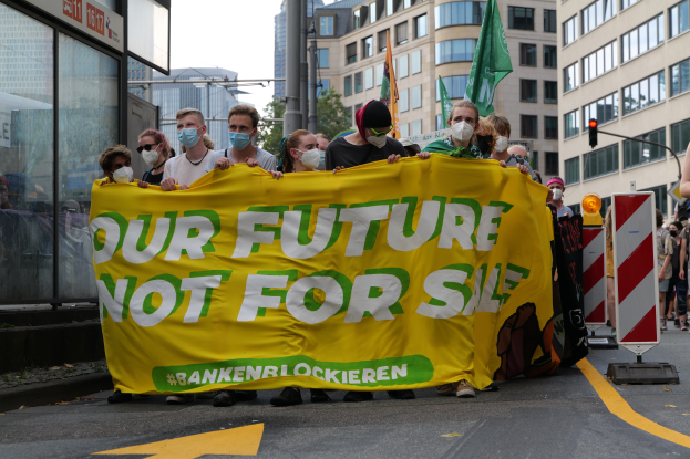 Eine Gruppe von Menschen marschiert mit Masken und einem gelben Banner mit der Aufschrift "Unsere Zukunft nicht verkaufen" die Straße entlang, mit Gebäuden, Laternenmasten, Ampeln, Bäumen und einem Schild im Hintergrund unter einem klaren blauen Himmel.
