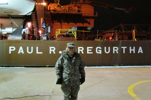 Eine Frau steht lachend, trägt eine Mütze und Handschuhe, mit einem Schiff und Text im Hintergrund.