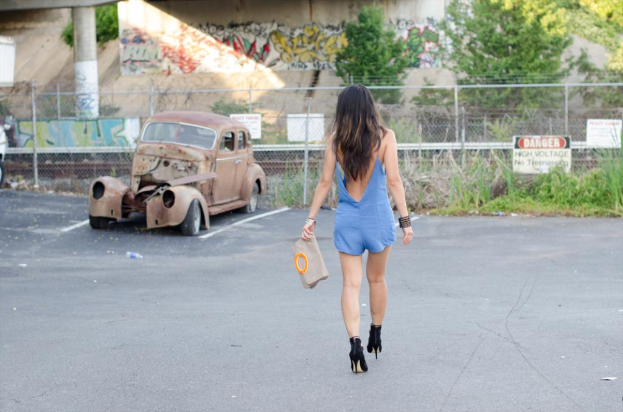 Eine Frau geht auf der Straße und hält eine Tasche in der Hand, mit einem geparkten Auto, Pflanzen, einem Gebäude und Bäumen im Hintergrund.