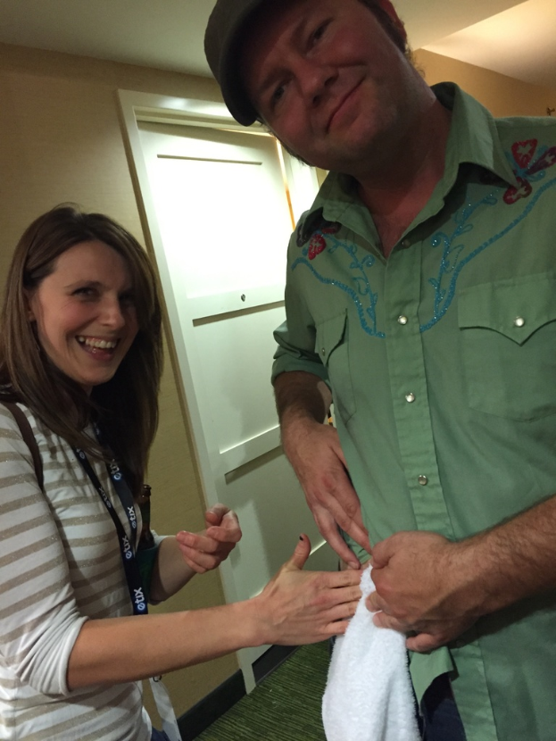 Ein Mann und eine Frau stehen und lächeln, mit einem weißen Handtuch zu ihren Füßen, einer Wand und einer weißen Tür hinter ihnen und einem Dach des Gebäudes darüber.