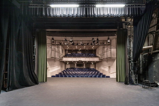 Ein großer Zuschauerraum mit schwarzen Vorhängen, einer Bühne, Deckenlampen und Stühlen, mit Gegenständen auf der rechten Seite, die auf einen Veranstaltungsort hinweisen.