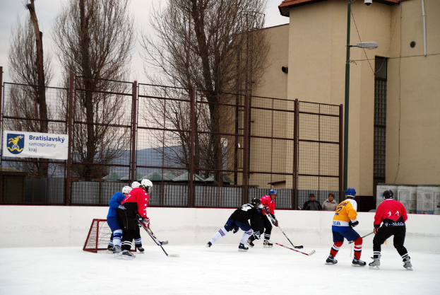 Menschen beim Eishockey-Spielen auf einer Eisfläche mit Gebäuden, Bäumen, einer Straßenlaterne, einem Namensschild und Zäunen im Hintergrund unter einem Himmel.