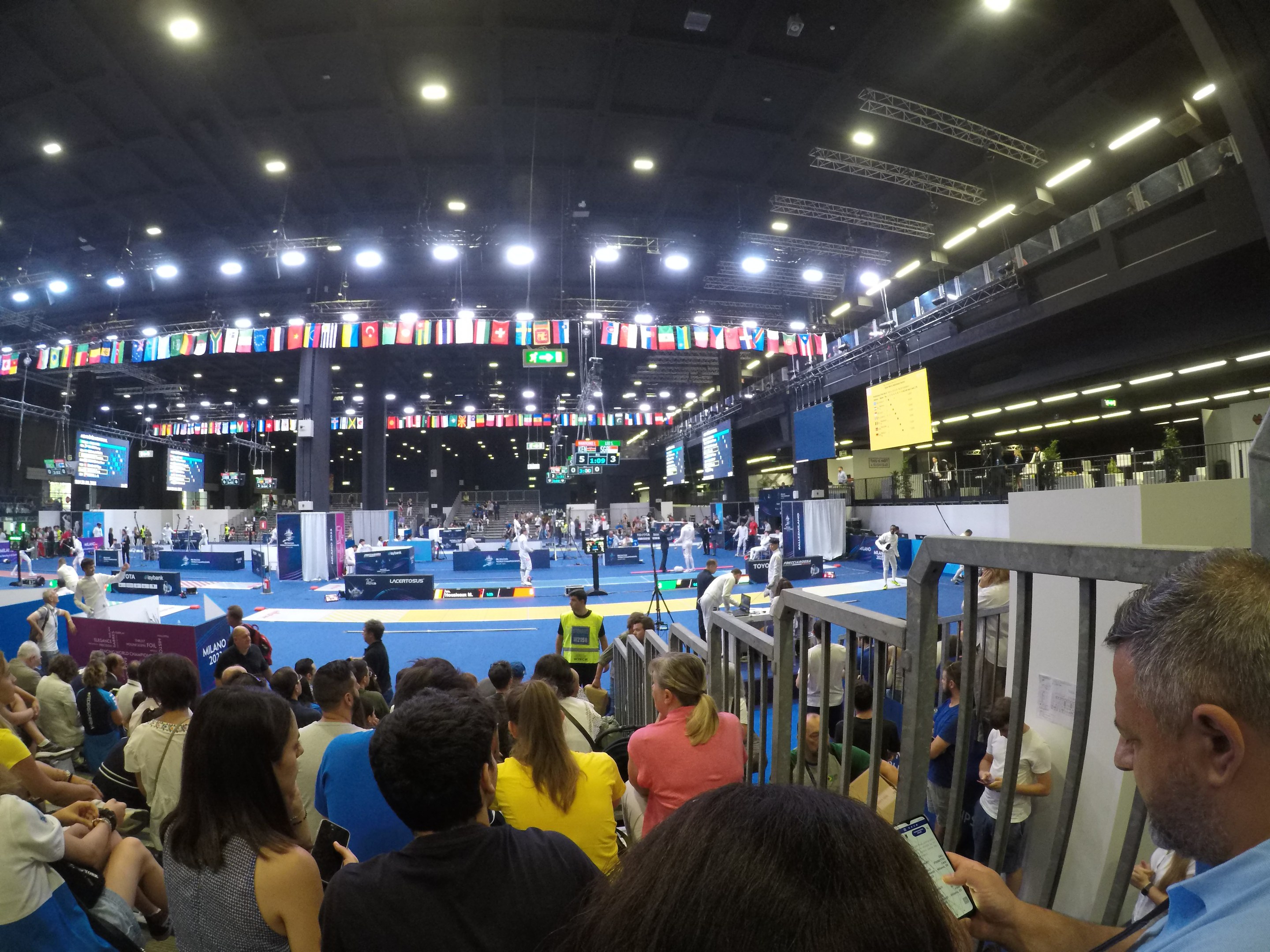 Eine große Menge von Menschen, die auf Sitzplätzen und Stehplätzen in einer Indoor-Sportarena sitzen, mit Bildschirmen, Flaggen und Deckenleuchten im Hintergrund.